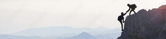 Silhouette zweier Bergsteiger vor Bergpanorama. Einer der Bergsteiger hilft beim Erklimmen eines Gipfels