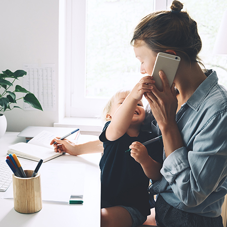 Eine junge Frau sitzt am Schreibtisch und notiert etwas. In der anderen Hand hält sie ein Smartphone, mit dem sie telefoniert. Das Kleinkind auf ihrem Schoß greift lachend nach dem Gerät.